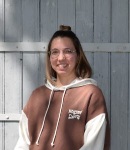 Das Bild zeigt eine junge Frau mit braun-blonden Haaren, einer runden Brille und einem brau-weißen Pullover. SIe steht vor einer grauen Brettertür.